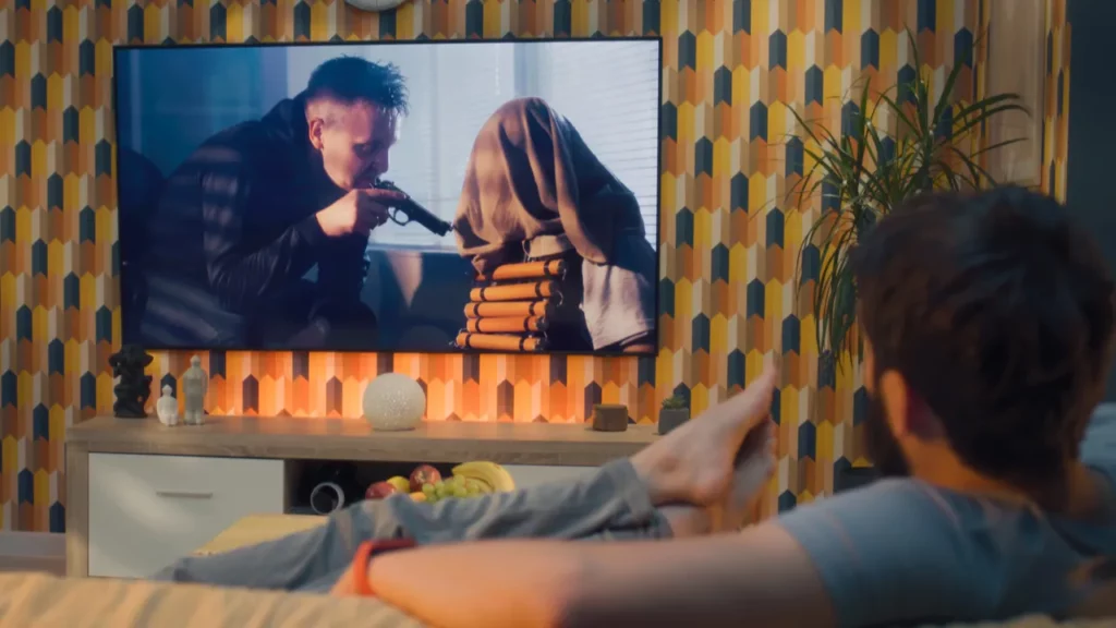 Asmuo, žiūrintis televizijos sceną su ginklu.